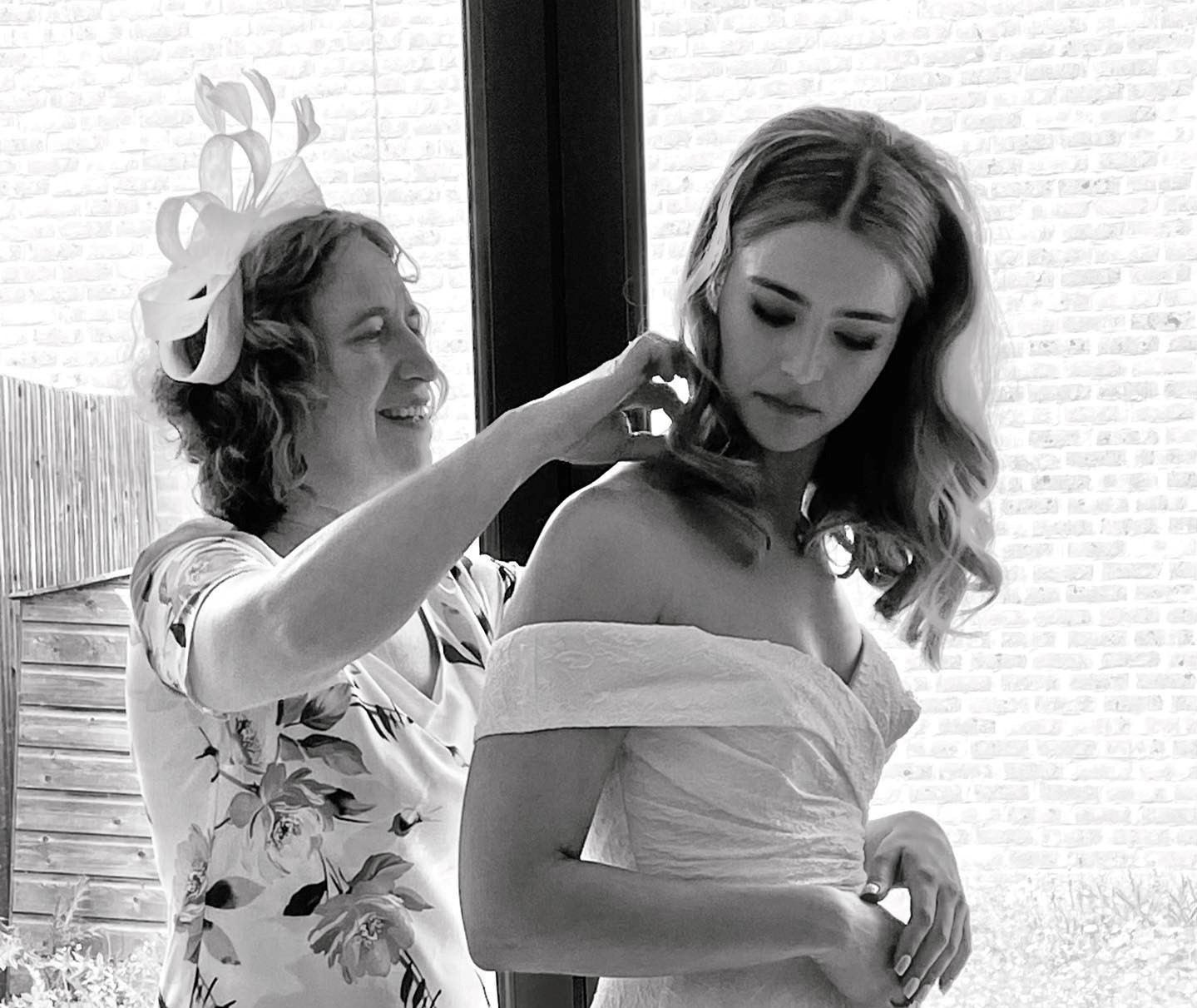 A black and white image shows Rachel, the translator, dressed in a floral print dress and a fancy hat as Mother of the Bride, helping her daughter, who is wearing a wedding dress, to fasten her necklace.