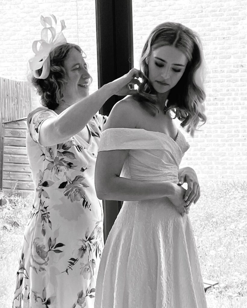 A black and white image shows Rachel, the translator, dressed in a floral print dress and a fancy hat as Mother of the Bride, helping her daughter, who is wearing a wedding dress, to fasten her necklace.